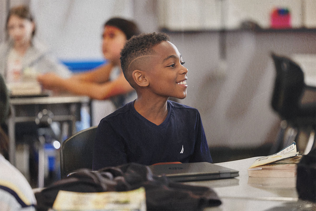 A student sits at a classroom desk, smiling and looking to the side.
